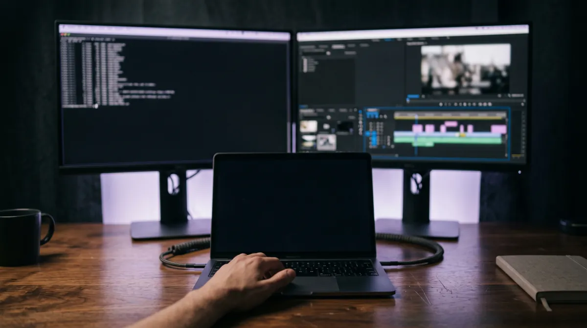 A desk at night with two displays, a terminal on the left and a video timeline on the right, above a laptop with a hand on its keyboard.