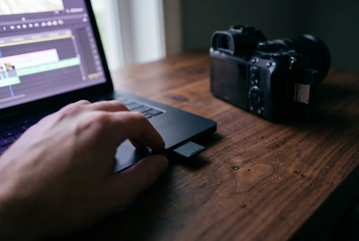 A hand slides a camera's SD card into the side of a MacBook Pro, the camera beside it with its card door open.